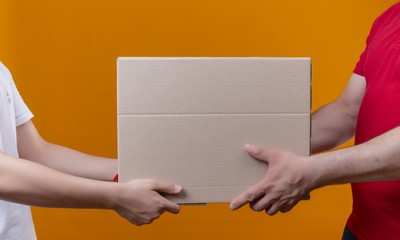 cropped view of delivery man in red uniform giving box package to a customer over isolated orange background